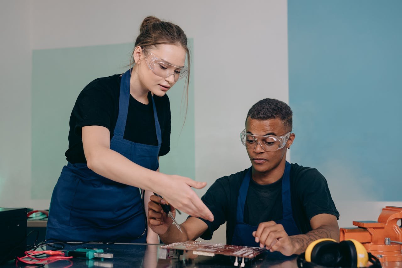Two engineers working together on electronic components, wearing safety goggles and aprons.
